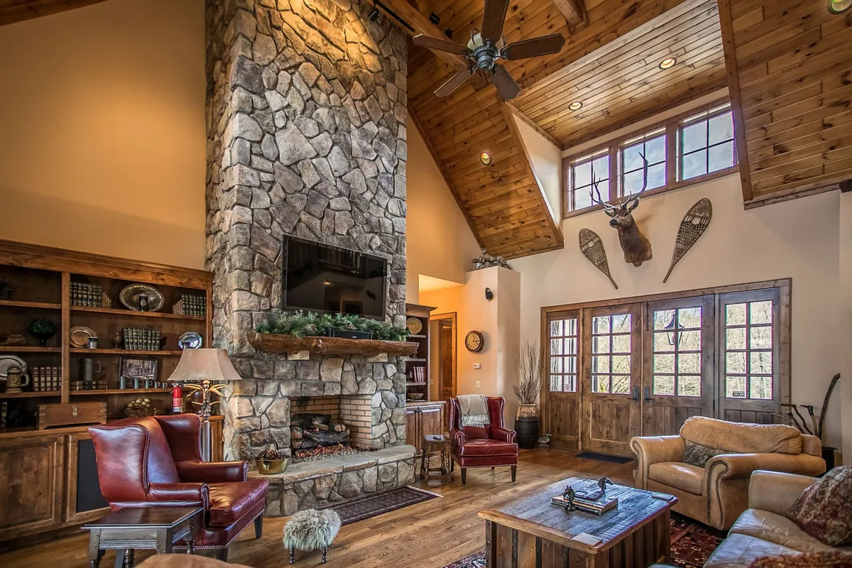 This image features a spacious great room highlighted by a grand stone fireplace, wooden bookshelves, large windows allowing in ample natural light, and a comfortable seating area arranged for socializing or relaxing by the fire. The rustic decor and architectural details evoke a sense of mountain luxury and warmth.