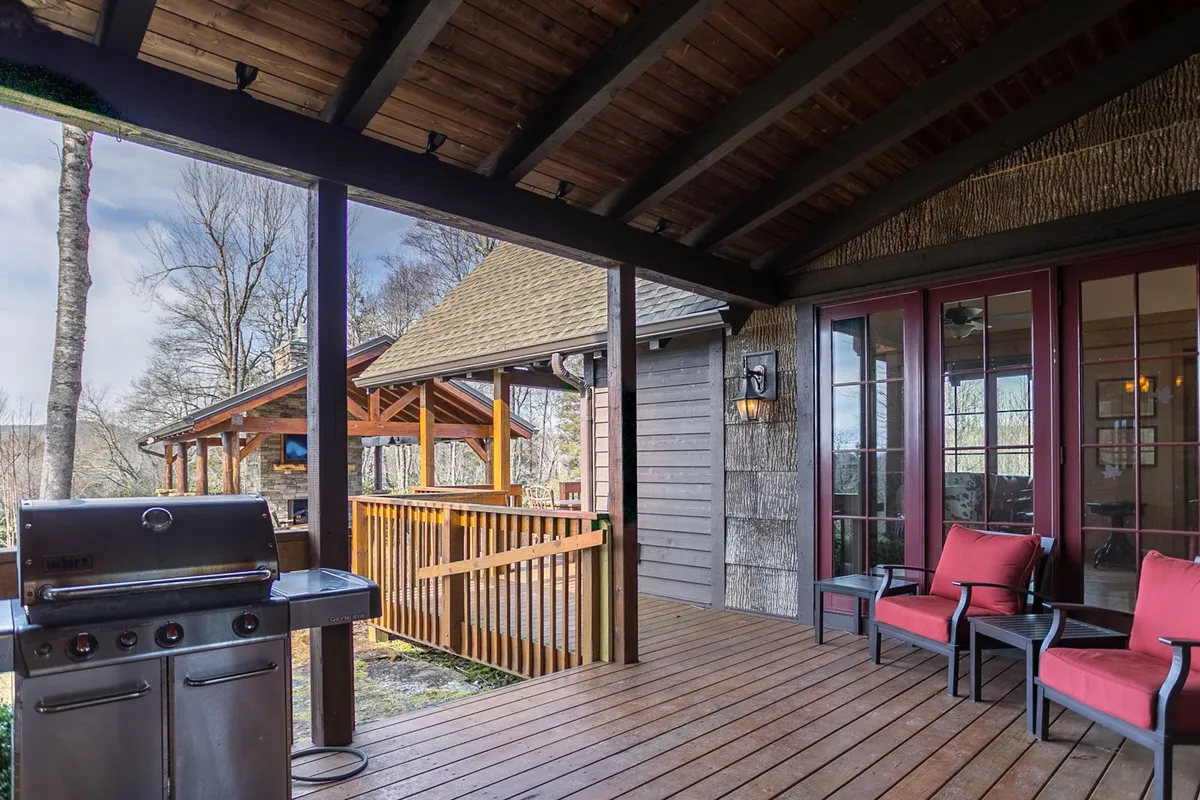The image shows a cozy wooden deck featuring a grill, red chairs, and lush trees in the background. The setup is ideal for cookouts and casual family gatherings, inviting guests to socialize, relax, and enjoy the outdoors together.