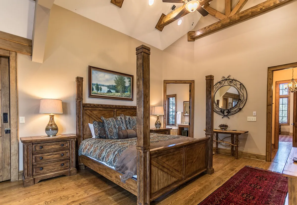 A four-poster bed sits against a wooden floor, accompanied by a nightstand with a lamp, a large mirror, and framed landscape art. Ample natural light streams through the window, creating a calm and cozy atmosphere, ideal for restful sleep and privacy.