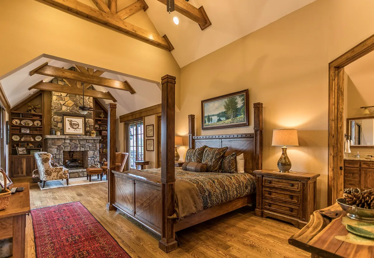 A spacious bedroom featuring a grand king-sized bed with an ornate headboard, a cozy stone fireplace, exposed wooden beams on the ceiling, and a comfortable seating area by large windows. The setting is luxurious yet welcoming, exuding warmth and privacy.