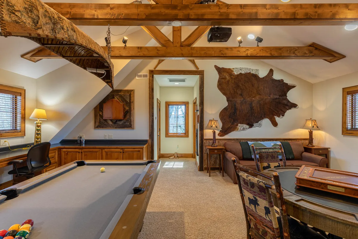 This inviting interior showcases a game room with a pool table at its center, surrounded by exposed wooden beams, a plush leather couch, and a decorative rug. The room exudes a welcoming atmosphere, ideal for gatherings and games, and gives a sense of rustic charm.