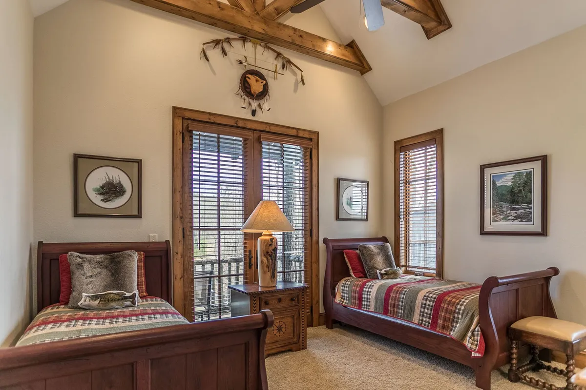 A rustic bedroom with two twin beds adorned in plaid bedding, a wooden bedside table with a lamp, decorative wall art, and large windows with wooden blinds. The space feels both functional and inviting, perfect for flexible sleeping arrangements or visiting children or friends.