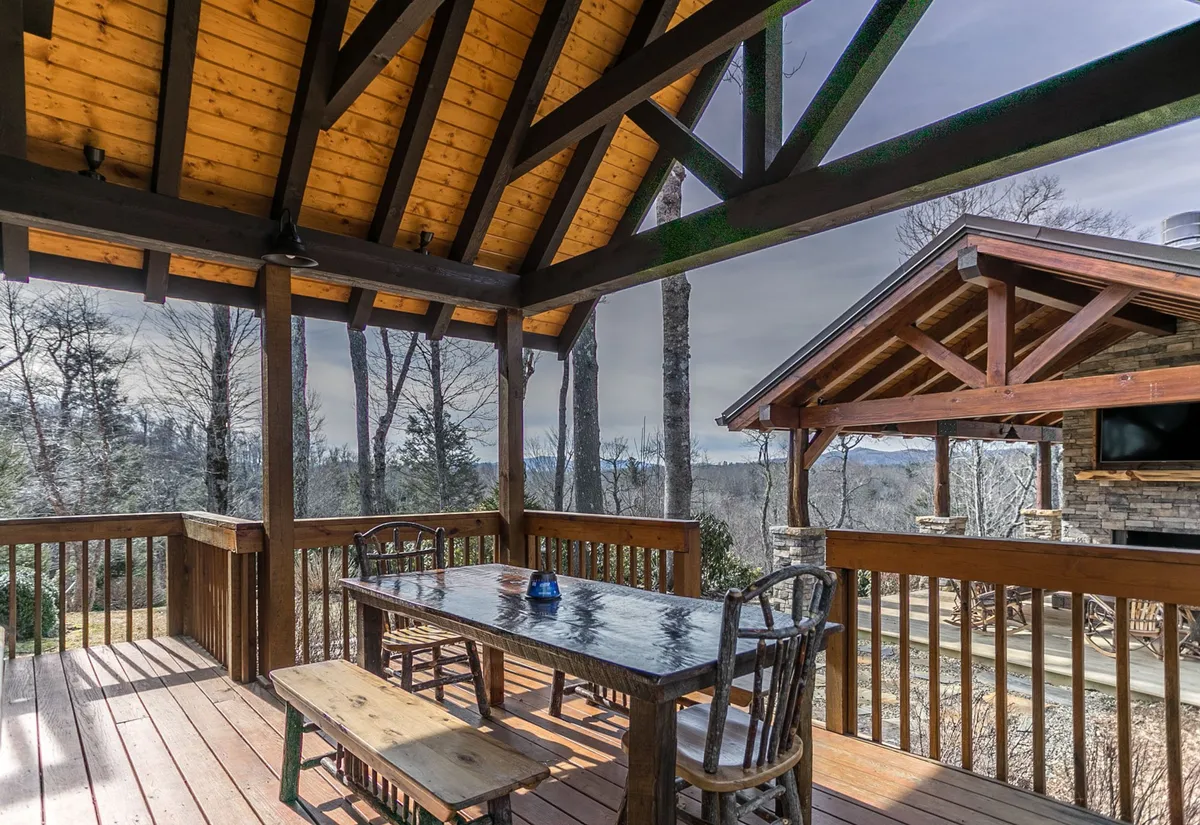 A spacious covered deck with a large wooden dining table, matching chairs, and exposed beams, all surrounded by trees. The setting suggests communal meals, outdoor relaxation, and seamless indoor/outdoor living.