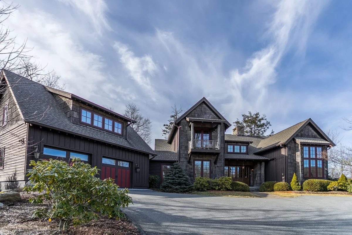 The image showcases the impressive stone facade of Elk Camp Lodge, with a grand entrance framed by striking red garage doors and a beautifully landscaped front yard. The setting is elegant yet inviting, hinting at the luxury and warmth awaiting inside, while the surrounding mountain landscape suggests tranquility and mountain charm.