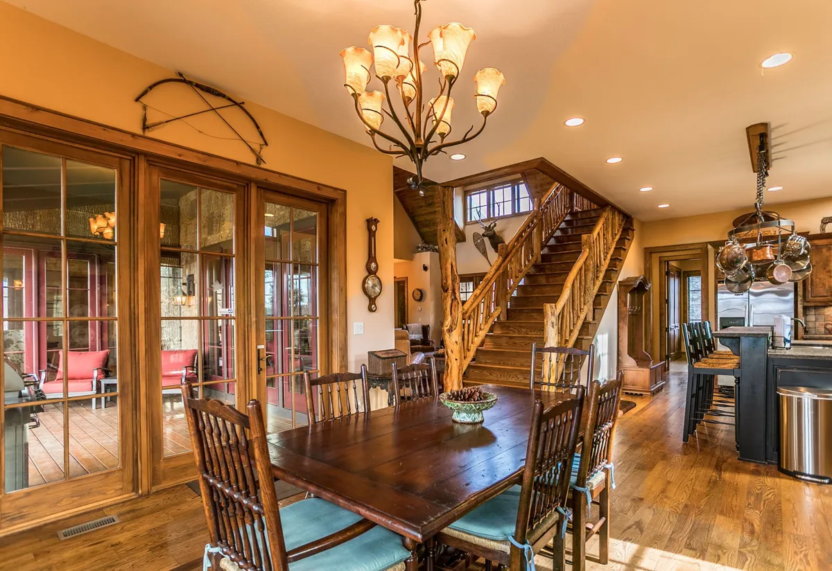 This interior scene highlights a dining area with a cherry table, chandelier, staircase, kitchen area, and large windows providing a view to the outdoors. The layout encourages socializing and showcases the open flow between dining and kitchen spaces, while natural light and rustic details enhance the ambiance.