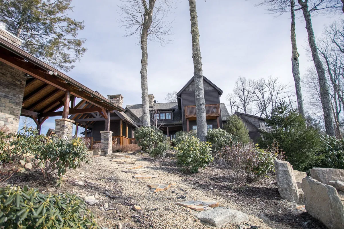 A charming stone pathway leads through large trees to a multi-story house surrounded by a landscaped garden. The outdoor area is generous and green, offering plenty of room for pets to roam and for guests to enjoy nature walks or gatherings.