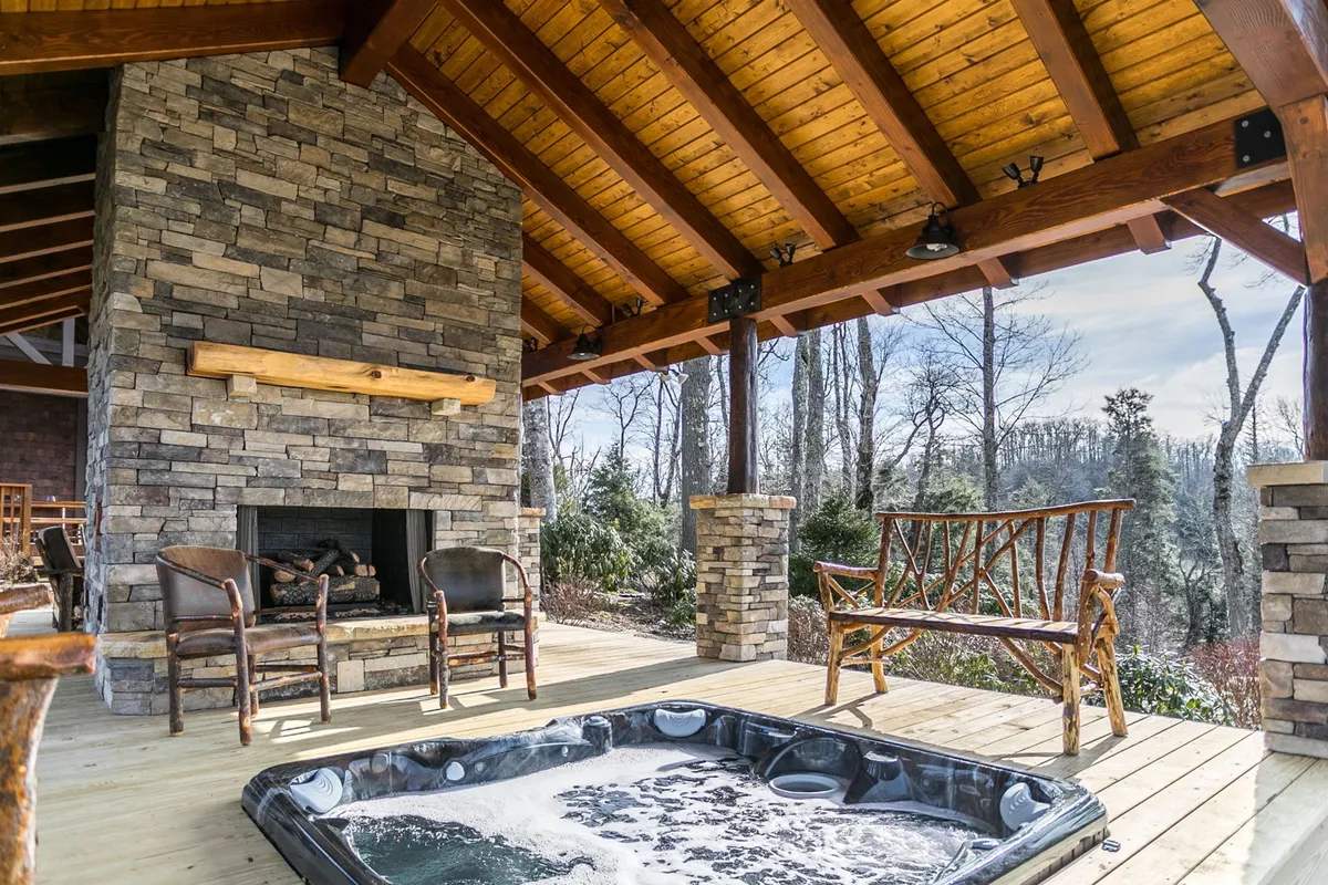 This image showcases an inviting outdoor living area with a stone fireplace, built-in hot tub, wooden seating, and a natural backdrop. The scene radiates warmth and comfort, perfect for both relaxation and entertaining guests under the open sky.