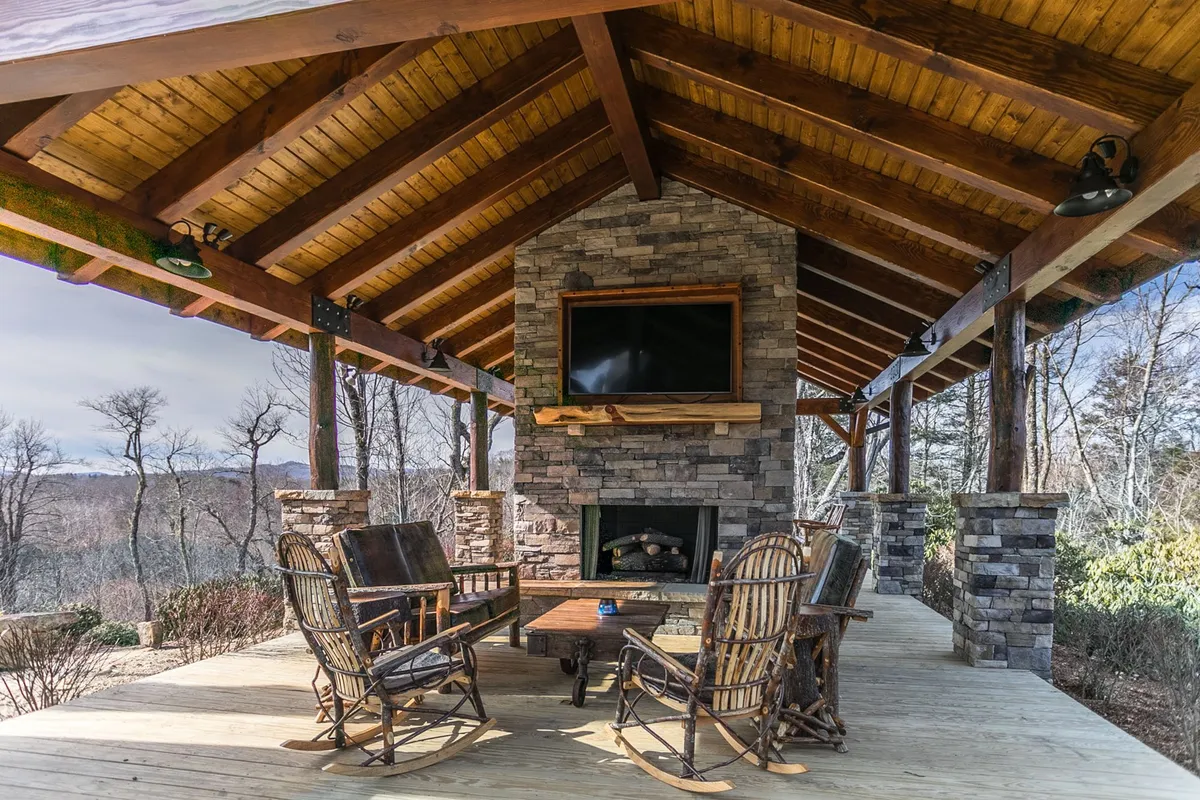 This outdoor pavilion features a striking stone fireplace, wooden rocking chairs, and a flat-screen TV, all set beneath robust wooden beams with views of the surrounding landscape. The space is designed for year-round entertaining and relaxation.